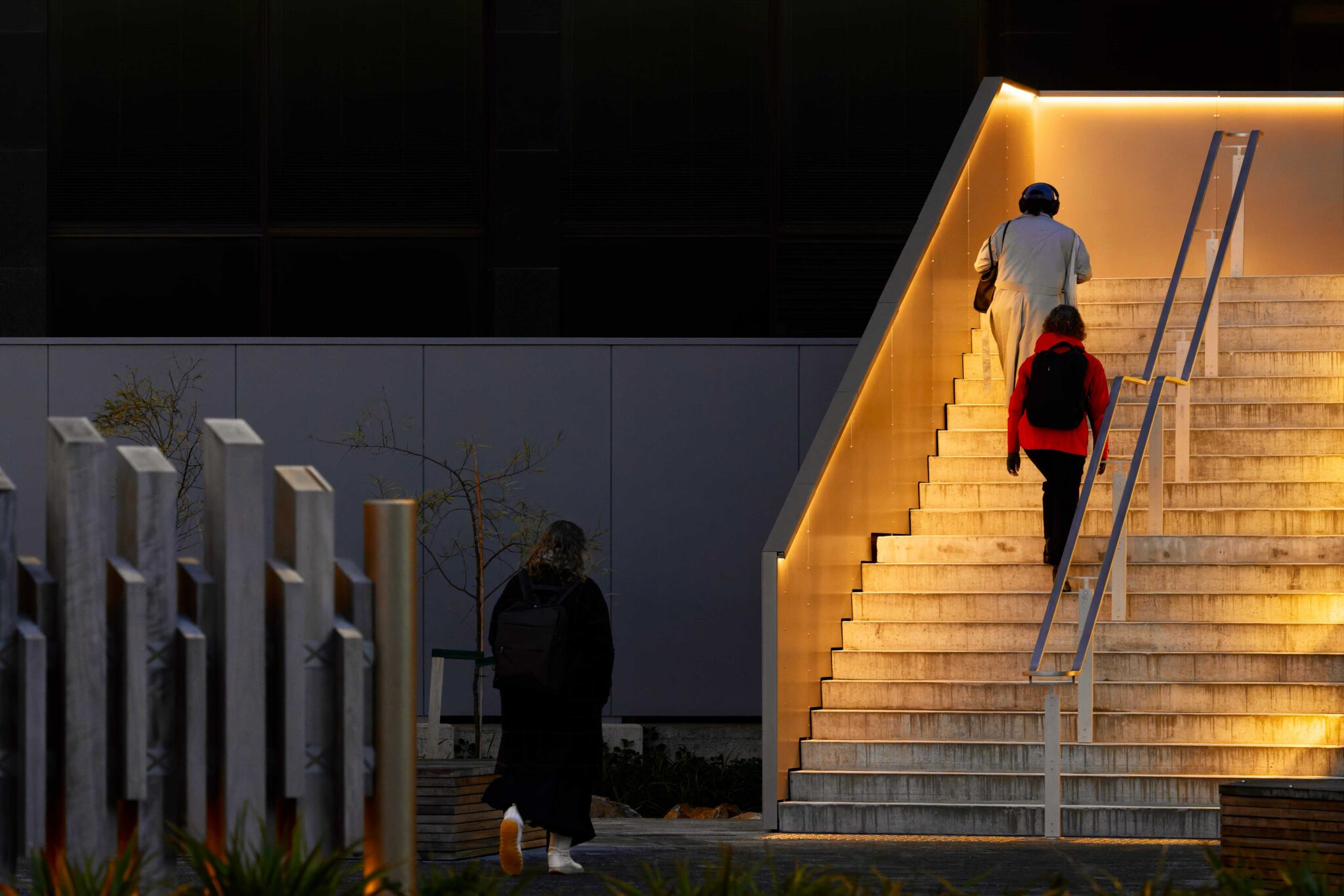 Exterior concrete staircase at Te Rua Archives New Zealand illuminated by Offspring Profiles’ Flat Freddie 30 integrated beneath the stair balustrade capping, with people walking up the steps at dusk.
